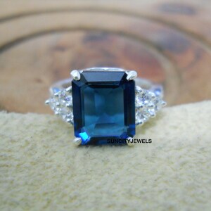 Può includere: Un anello d'argento con una grande gemma rettangolare blu scuro come elemento centrale. Gemme più piccole e chiare affiancano la pietra principale. L'anello è posto su uno sfondo neutro. Il testo "SUNCITYJEWELS" è visibile.