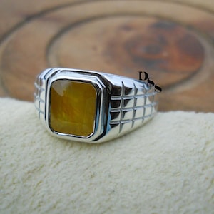 Peut inclure: Une bague en argent avec une pierre précieuse jaune rectangulaire. La bague a un motif texturé sur le bandeau. La pierre précieuse est d'une couleur jaune doré chaud, sertie dans un sertissage en argent poli. La bague est présentée sur une surface crème.