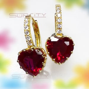 Puede incluir: Un par de pendientes de aro dorados con piedras preciosas rojas en forma de corazón. Los aros están adornados con pequeñas piedras preciosas transparentes. Los pendientes se muestran sobre un fondo suave y colorido.