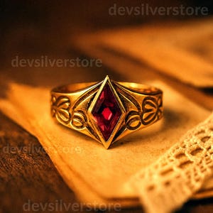 May include: A gold ring featuring a central red, diamond-shaped gemstone. The ring has an ornate design with floral patterns on the band. The ring is displayed on aged paper.