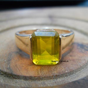 Peut inclure: Une bague dorée avec une grosse pierre jaune rectangulaire. Le chaton est lisse et poli, et la pierre est facettée. La bague est posée sur une surface en bois.