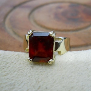 May include: A gold ring featuring a large, rectangular, dark red gemstone. The gemstone is set in a gold band with four prongs holding it in place. The ring is displayed on a cream-colored surface.