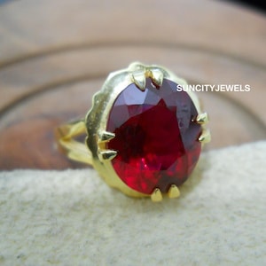 Könnte beinhalten: Ein goldener Ring mit einem großen, oval geschliffenen, tiefroten Edelstein. Der Edelstein ist in einer goldenen Lünette mit gewelltem Rand und kleinen dekorativen Zinken gefasst. Der Text "SUNCITYJEWELS" ist sichtbar.