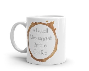 May include: White ceramic coffee mug with a brown coffee stain design. The mug features the text "A Bissell Meshuggah Before Coffee" in a black, sans-serif font. The mug has a white handle.