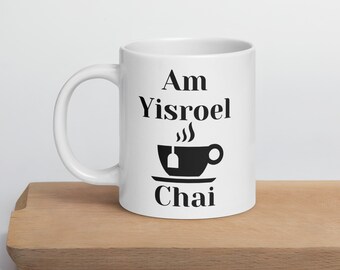 May include: White ceramic mug with the text "Am Yisroel Chai" in black. The design includes a stylized teacup with a tea bag and steam. The mug is on a wooden surface.