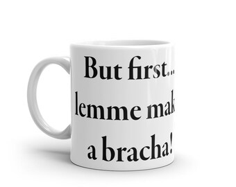 May include: White ceramic coffee mug with the text "But first... lemme make a bracha!" in black, bold font. The mug has a curved handle and a glossy finish. The mug is designed for hot beverages.