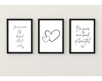 Op de afbeelding: Drie zwarte ingelijste prints met witte achtergronden en zwarte tekst. De linker print zegt "You're never fully dressed without a smile." De middelste print heeft twee zwarte harten. De rechter print zegt "Be your own kind of beautiful."