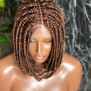 May include: A bob-style wig featuring tightly braided strands in shades of brown and light brown. The braids are neatly arranged, creating a symmetrical pattern. The wig is styled on a mannequin head, showcasing the hairline and overall design.
