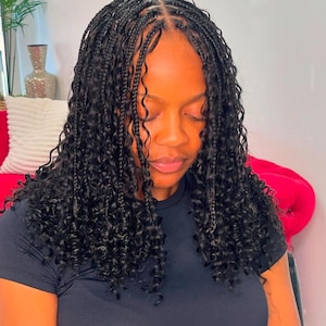May include: A person with long, black, curly braids. The braids are styled with small, neat sections at the crown of the head, transitioning into longer, flowing curls. The person is wearing a black t-shirt.