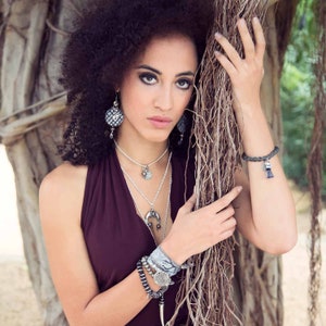 May include: A woman with curly dark hair wearing a burgundy halter top and silver jewelry, including a layered necklace and bracelets. She is leaning against a tree trunk with vines.
