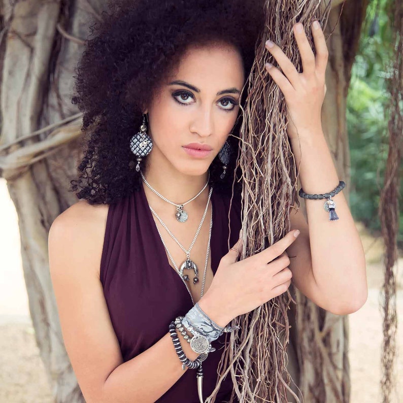 May include: A woman with curly dark hair wearing a burgundy halter top and silver jewelry, including a layered necklace and bracelets. She is leaning against a tree trunk with vines.