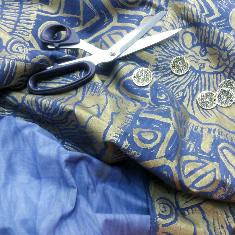 May include: A blue fabric with gold metallic designs and a pair of silver scissors. There are several gold metal circular embellishments on the fabric.