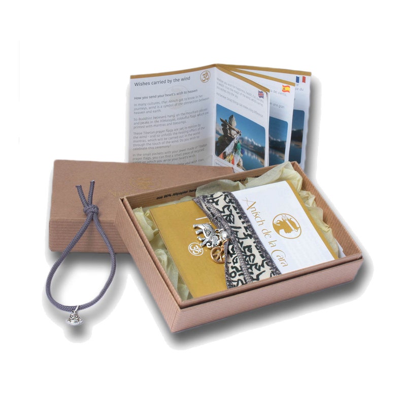 May include: A small gift box with a brown lid and a gray ribbon. Inside the box is a small yellow book, a gray and white patterned ribbon with charms, and a white card with the text "Anuschka de la Cara".