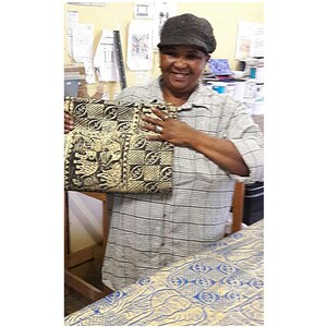 May include: A woman holds a black and yellow tote bag with a geometric pattern. The bag is made of fabric and has a woven texture. The woman is wearing a plaid shirt and a gray hat.
