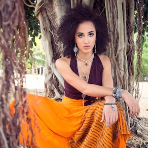 May include: A woman with curly dark hair wearing a brown halter top and an orange and brown patterned skirt. She is sitting under a tree and wearing silver jewelry, including earrings, necklaces, and bracelets.