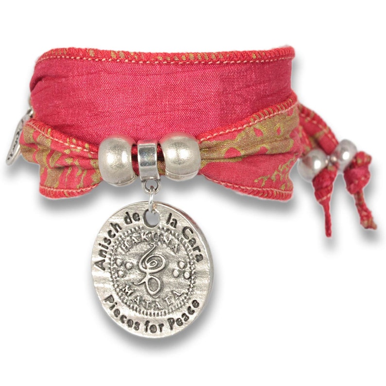 May include: A red and gold fabric bracelet with a silver charm that says "Anisch de la Cara" and "Piecos for Peace".