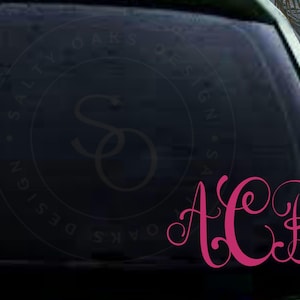 May include: A pink monogram decal with the letters ACB on a black car window. The decal is surrounded by a circle with the text "Salty Oaks Design" in a white font.