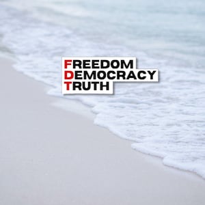 May include: A white rectangular sticker with the words "FREEDOM DEMOCRACY TRUTH" in black and red text, placed on a sandy beach with ocean waves in the background.