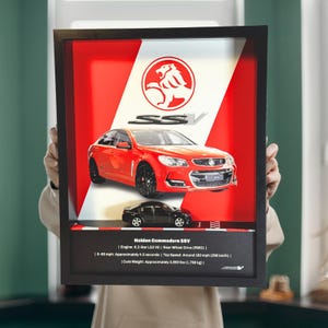 May include: Framed artwork featuring a red Holden Commodore SSV car model. The background has a red and white design with the SSV logo. The frame also includes a smaller black car model and text with car specifications.