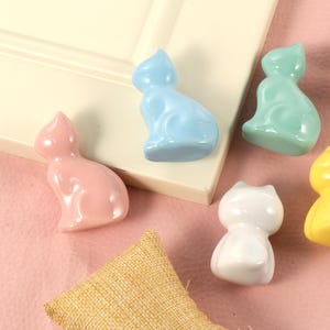 Puede incluir: Seis figuras de gato de cerámica en tonos pastel de rosa, azul claro, verde menta, blanco y amarillo. Las figuras están dispuestas sobre una superficie blanca y un fondo rosa, con un pequeño saco de yute en primer plano.