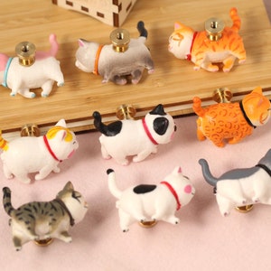 Può includere: Una collezione di nove pomelli per mobili a forma di gatto. Ogni pomello presenta un gatto di colore diverso con una vite in ottone. I gatti sono bianchi, arancioni, grigi e neri, con collari di vari colori. I pomelli sono su una superficie di legno.