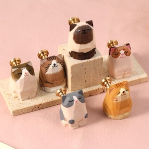 May include: A collection of wooden cat figurines with gold-colored tops. The cats are in various colors and patterns, including brown, white, gray, and orange. They are arranged on small, light-colored platforms against a pink background.