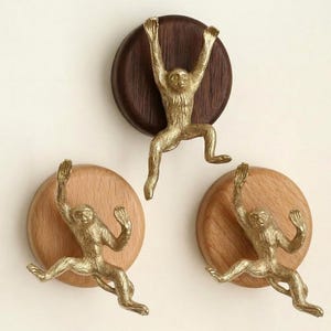 May include: Three decorative wall hooks with brass monkey figures. Each monkey is posed climbing, attached to a round wooden base. The bases are in shades of brown and natural wood.