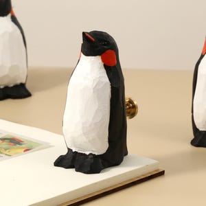 Peut inclure: Trois figurines de pingouins en bois sculpté, avec des détails noirs, blancs et rouges. L'un des pingouins est fixé à un bouton de tiroir. Les pingouins sont sur une surface claire, avec un livre ouvert au premier plan.