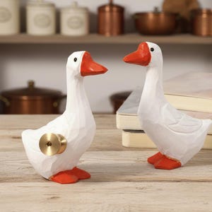 Op de afbeelding: Twee witte ganzenbeeldjes met oranje snavels en poten. Eén gans heeft een goudkleurige knop aan de zijkant. De ganzen staan op een houten oppervlak, met boeken en keukengerei op de achtergrond.