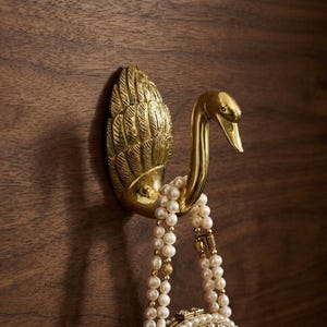 Könnte beinhalten: Ein messingfarbener, schwanförmiger Wandhaken mit detailliertem Feder- und Flügeldesign. Der Haken hält eine Perlenkette und eine kleine, verzierte Handtasche. Der Haken ist auf einer dunklen Holzoberfläche montiert.