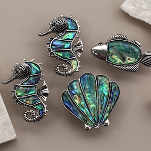 May include: A collection of silver-toned sea-themed jewelry pieces. The set includes two seahorse brooches, a fish brooch, and a seashell brooch. Each piece features iridescent abalone shell inlays, showcasing shades of green, blue, and gold.
