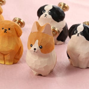 Puede incluir: Figuras de perros de madera en varios colores, incluyendo naranja, negro y blanco. Cada figura tiene un pomo de color latón. Los perros tallados se exhiben sobre una superficie rosa. Las figuras miden unos 5-7 cm de alto.