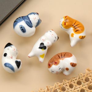 Può includere: Cinque statuette di gatto in ceramica in varie pose. I gatti sono bianchi con segni blu, arancioni e marroni. Un gatto ha un cuore nero. Le statuette sono su una superficie beige chiaro.