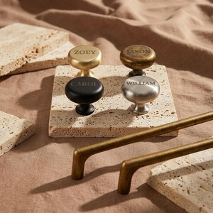 Puede incluir: Cuatro pomos de armario en oro, bronce, negro y plata, cada uno con un nombre grabado: Zoey, Jason, Carol y William. Dos tiradores de armario de color bronce están en primer plano. Los pomos y tiradores se muestran sobre una superficie clara y texturizada.