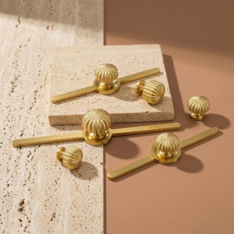 May include: A selection of gold-toned cabinet hardware, featuring ribbed knobs and rectangular pulls with central knobs. The knobs have a pumpkin-like design. The hardware is presented on a textured, light-coloured surface.