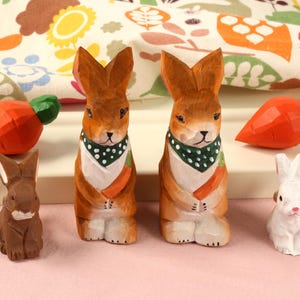May include: Wooden rabbit figurines and carved carrots. Two larger brown rabbits wear green and white polka dot scarves and hold carrots. A small brown rabbit and a small white rabbit are also present. The carrots are orange with green tops.