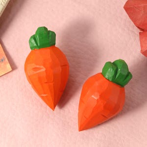 Op de afbeelding: Twee gesneden houten wortels met groene toppen en oranje lichamen worden gepresenteerd op een roze oppervlak. De wortels hebben een facetontwerp, waardoor ze een geometrische uitstraling hebben. De achtergrond omvat een wit notitieboek en een rode papieren waaier.