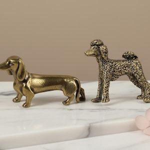 May include: Two brass dog figurines, a dachshund and a poodle, are displayed on a white marble surface. The dachshund is lying down, while the poodle is standing. The figurines have intricate details and a vintage aesthetic.