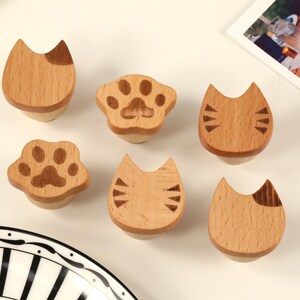 Puede incluir: Seis tiradores de cajón de madera con temática de gato con diferentes diseños, que incluyen una huella de pata, una cara de gato y una cola de gato. Los tiradores están hechos de madera marrón claro y tienen un acabado natural.