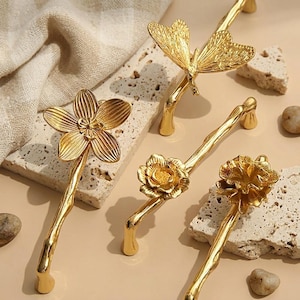 May include: Close-up of four gold-colored cabinet pulls with nature-inspired designs. One features a flower, another a butterfly, and two others have floral motifs. The pulls are displayed on a textured, light-colored surface.