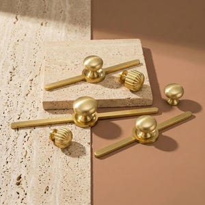 May include: A selection of gold-coloured cabinet hardware, including knobs and pulls. The knobs are round, while the pulls have a bar design. Some knobs have a ribbed texture. The hardware is displayed on a neutral background.