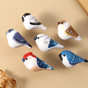 May include: Seven hand-painted ceramic bird figurines in various colours. The birds feature white, blue, red, and brown plumage with black accents. They are arranged on a beige surface.