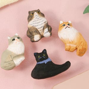 May include: Four hand-carved wooden cat figurines in various poses. The cats are painted in shades of gray, brown, orange, and black, with details like eyes and markings. A black cat wears a blue bow tie.