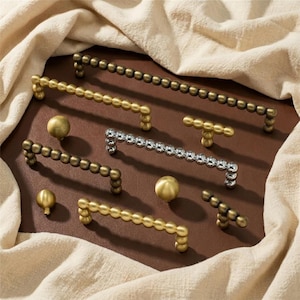 May include: A collection of decorative cabinet hardware, including handles and knobs. The handles are in various finishes, including gold, silver, and bronze. The knobs are spherical. The hardware is arranged on a brown surface with a beige fabric.