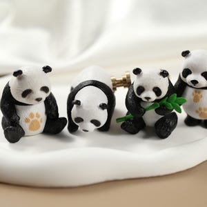 Peut inclure: Quatre petites figurines de panda noir et blanc. Les pandas sont debout sur une surface blanche. Un panda tient une feuille verte.