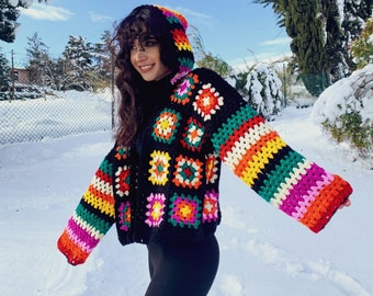Puede incluir: Una chaqueta de ganchillo negra con un diseño de patchwork colorido. La chaqueta tiene una capucha y mangas largas con vibrantes rayas arcoíris. La chaqueta se usa en un entorno nevado al aire libre.