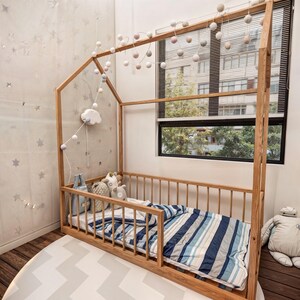 May include: A wooden house-shaped bed frame with a blue and white striped comforter. The bed is decorated with a string of white pom-poms and a cloud-shaped nightlight.