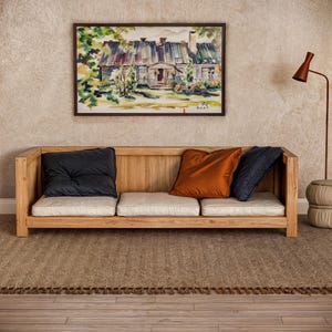 Op de afbeelding: Een houten bank met een lichtgekleurd kussen en drie kussens. De bank staat voor een beige muur met een ingelijst aquarel schilderij van een huis. De vloer is bedekt met een bruin geweven tapijt.
