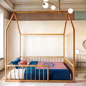 May include: A wooden toddler bed with a house-shaped frame. The bed has a blue and red striped mattress with a white design. The bed is in a room with a white wall and a wooden floor.