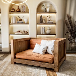 Puede incluir: Un gran sillón de madera con un cojín marrón y dos almohadas blancas. El sillón está en una habitación con una alfombra marrón claro y paredes blancas. Hay estantes con libros y objetos decorativos en las paredes.